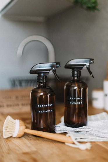 Two labeled spray bottles for bathroom and kitchen cleaning with a wooden scrubbing brush on a wooden surface.
