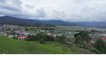 Cloudy sky over a green hilly area with scattered buildings.