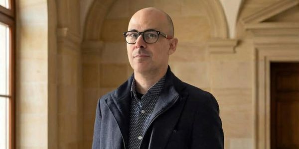 A man in glasses and a dark coat stands indoors with an arched stone background.