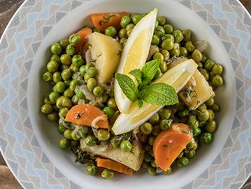 A bowl of cooked peas, carrots, and potatoes garnished with lemon wedges and mint leaves.