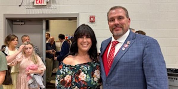Two people posing and smiling at an indoor event with others in the background.