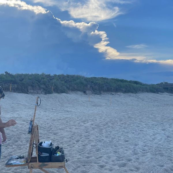 A man painting on a beach under a cloudy sky.