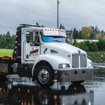 White flatbed tow truck parked on wet pavement with green trees in background.