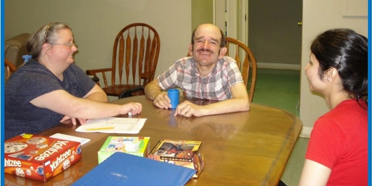 A SDS client is smiling at a table with two direct support workers as they play Yahtzee.