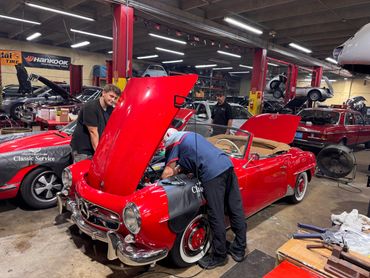 Two mechanics work on a red classic Mercedes convertible in a busy auto repair shop.