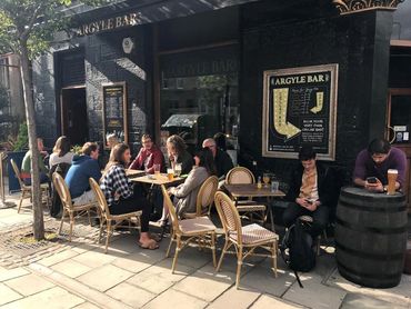 People are sitting and drinking on a sunny day in the outdoor seating area of the Argyle Pub and Cel