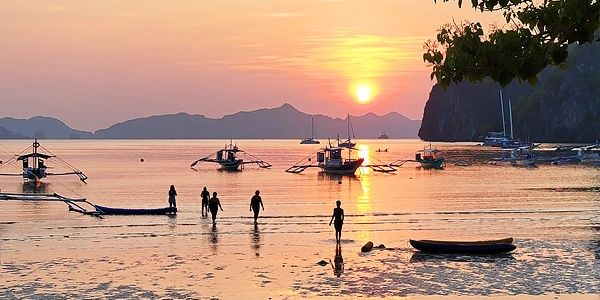 Sunset with people and boats on a calm beach with mountains in the background.