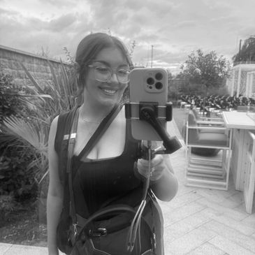 Woman smiling taking a selfie outdoors with a phone on a selfie stick.