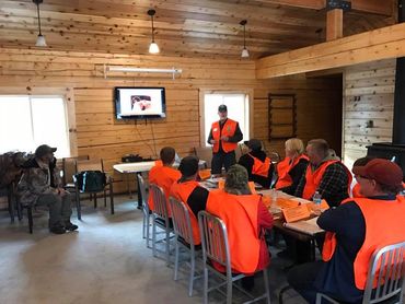 A group in orange safety vests attends a presentation inside a wooden room.