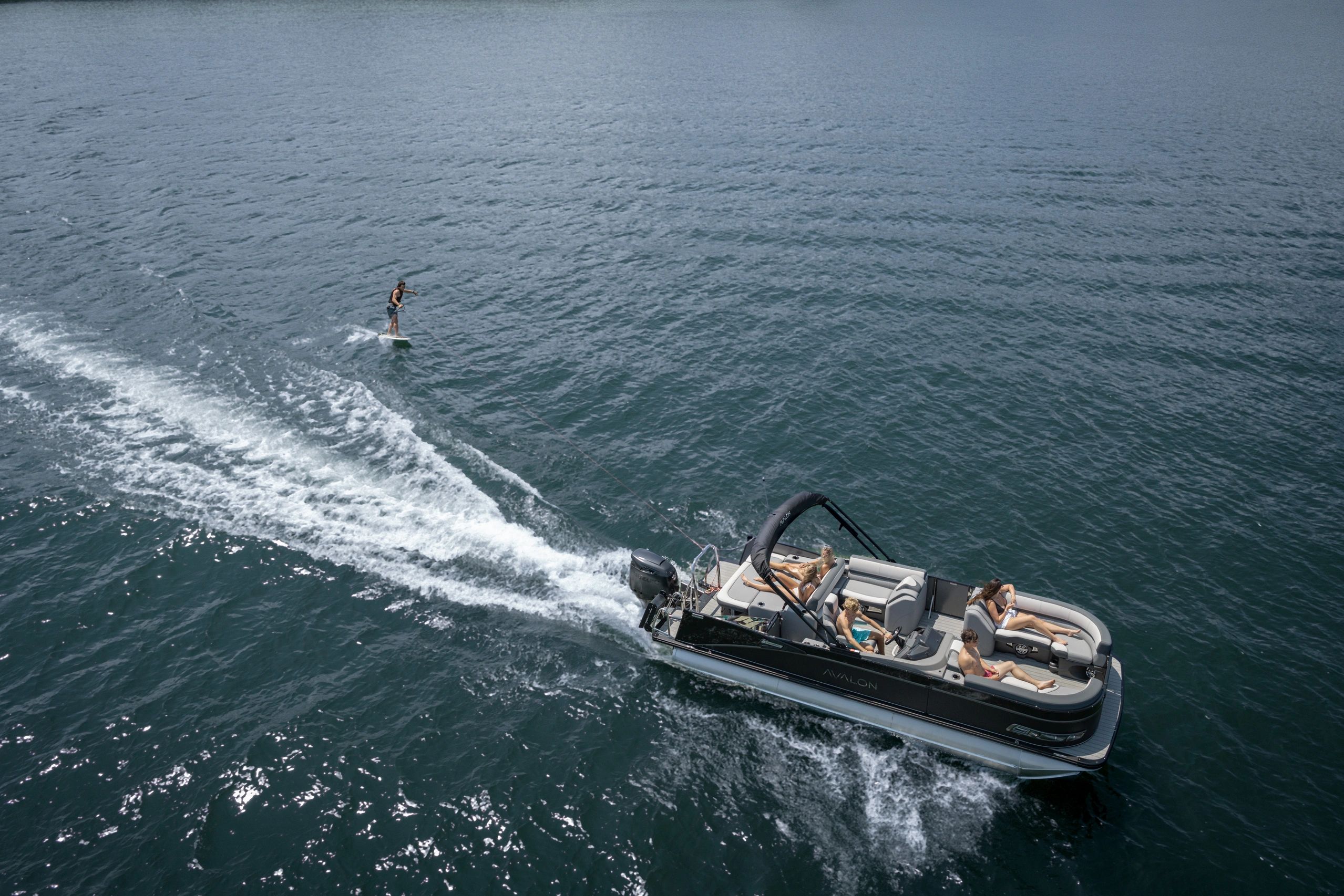 Person wakesurfing behind a pontoon boat with friends onboard.