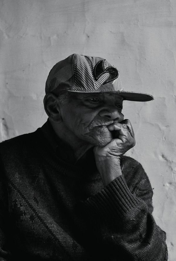 Elderly black man with baseball cap, sitting in a pensive mood, chin in hand, B/W image
