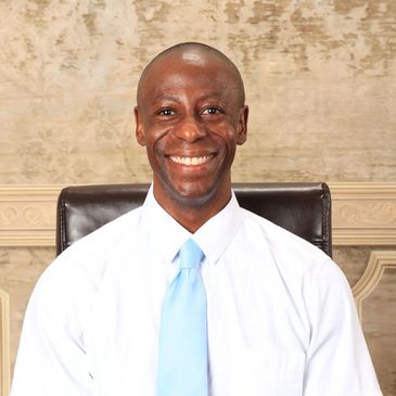 Smiling man in white shirt and light blue tie sitting on a chair.
