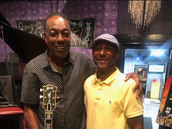 Two men smiling, one holding a guitar, inside a music-themed room.