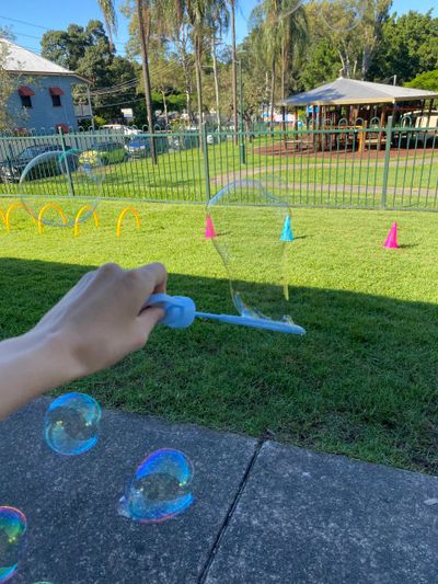 Hand blowing bubbles in a sunny park with colorful cones and hoops. Neurodiverse Playhouse