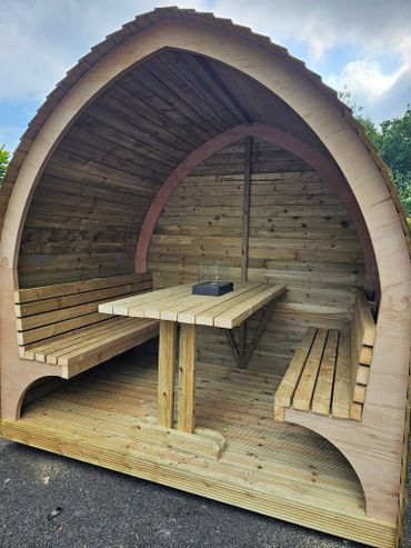 Wooden outdoor seating pod with benches and a table under a curved roof.