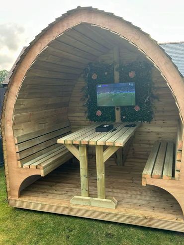Wooden outdoor pod with benches, table, and TV for watching sports.