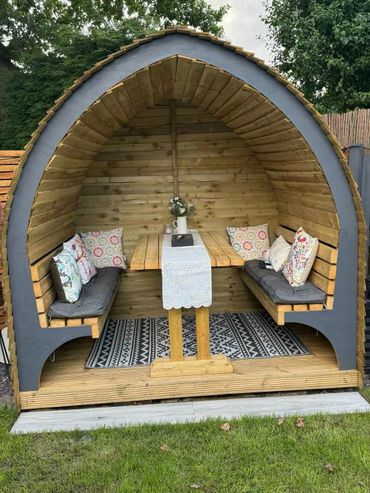Wooden garden pod with cushioned seating and a table inside.
