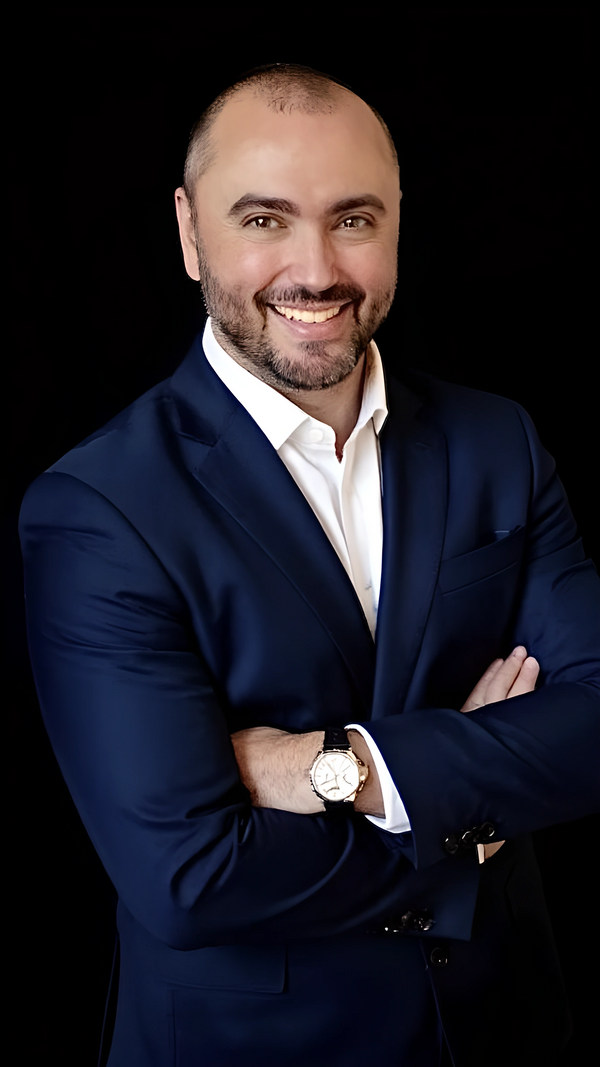 Smiling man in a navy suit with crossed arms against a black background.