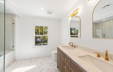 Bright modern bathroom with double sinks, gold fixtures, and marble floors.