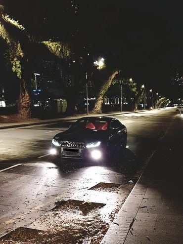 A black Audi with bright headlights parked on a quiet, tree-lined street at night.