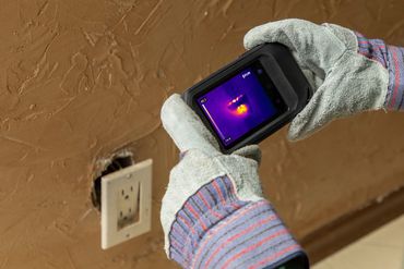 Gloved hands holding a FLIR thermal infrared camera checking an open electrical receptacle
