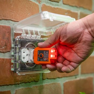 Hand holding a GFCI tool testing an outdoor bubble receptacle on a brick wall