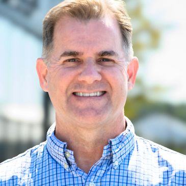 Smiling man in a blue checkered shirt outdoors on a sunny day.
