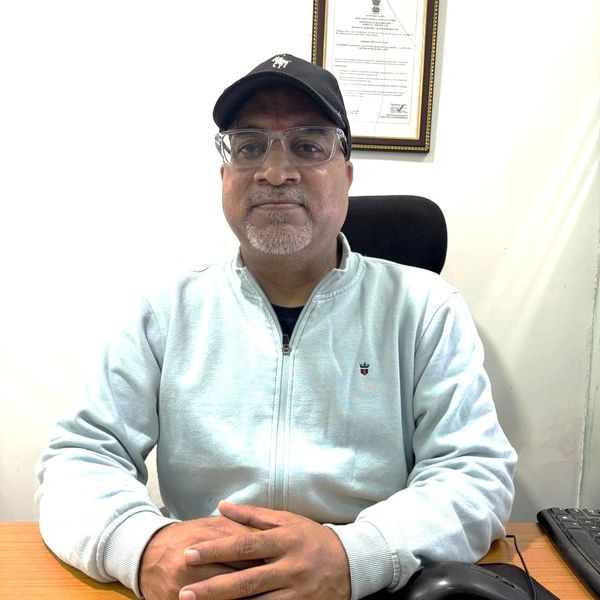 Man wearing glasses and cap sitting at a desk with hands folded.