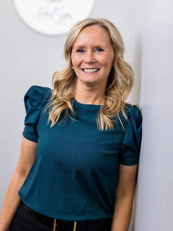 Smiling woman with blonde hair in a teal top leaning against a white wall.
