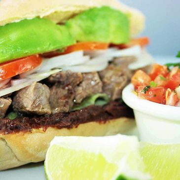 Close-up of a sandwich with beef, avocado, tomato, and onions, served with lime and salsa.