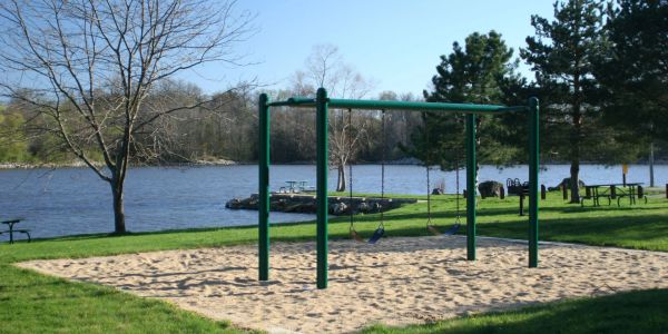 Empty swings in a sandy playground by a peaceful lake.