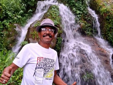 Man posing happily with thumbs up in front of a lush waterfall.