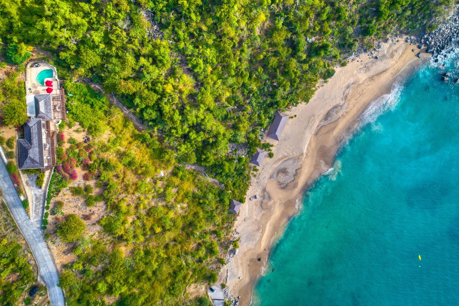 Aerial view of a villa with pool next to a lush green forest and a turquoise beach.