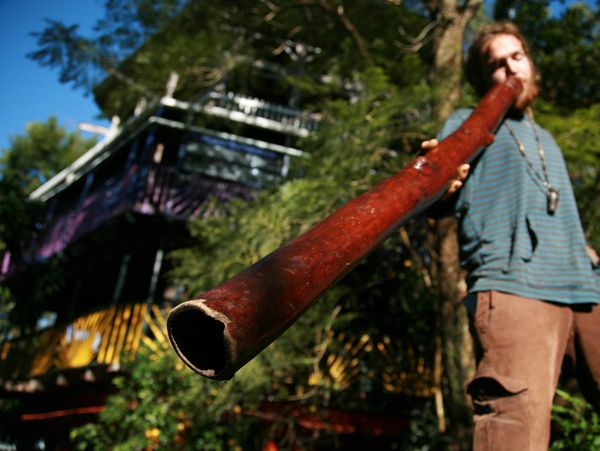 Man playing a large didgeridoo outdoors near a colorful building.