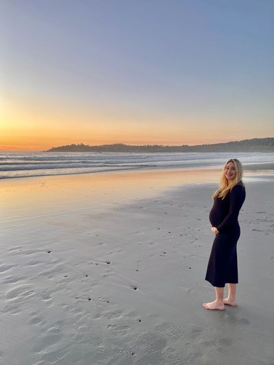Pregnant woman in black dress standing barefoot on the beach at sunset.