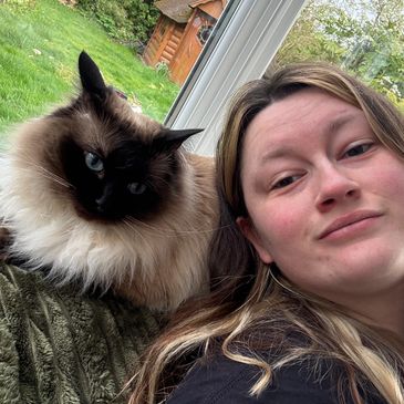 A woman takes a selfie with a fluffy Siamese cat resting on her shoulder.