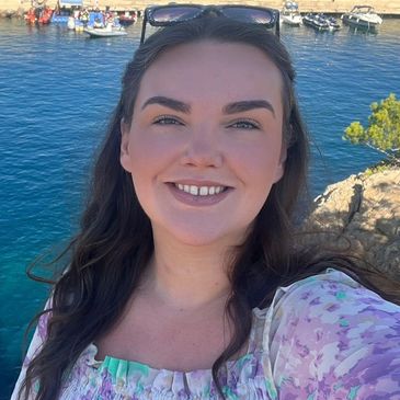 A smiling woman taking a selfie near a marina with boats and clear blue water.