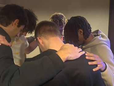 A group of young men in a huddle with heads bowed and hands on shoulders.