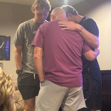 Three men in a close, supportive group hug indoors.