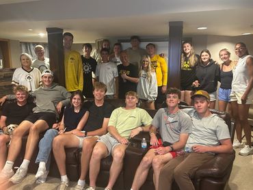 A large group of young adults and a small dog pose in a cozy living room.
