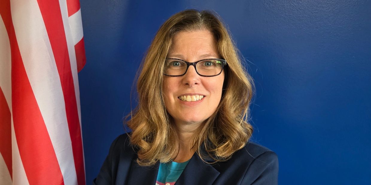 Smiling woman with glasses in front of an American flag and blue wall.