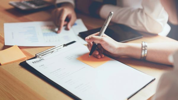 A person holding a pen over a resume on a clipboard during a meeting.