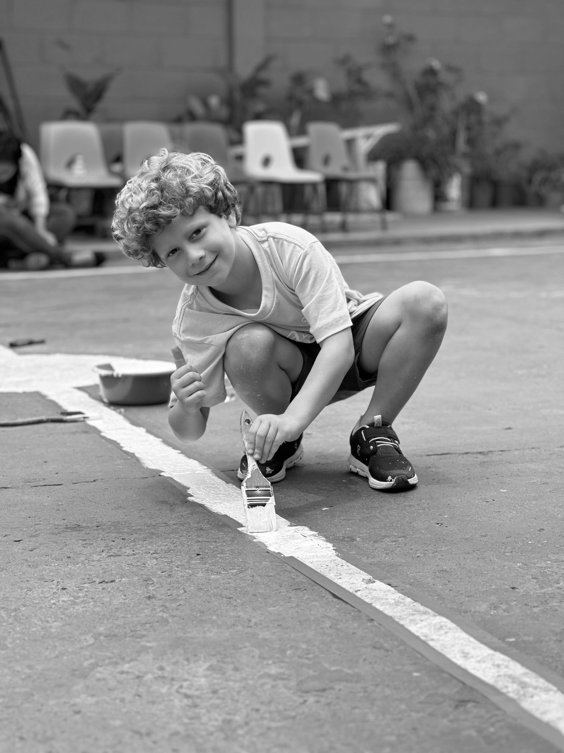 A young boy smiles and gives a thumbs up while painting a white line on the ground.