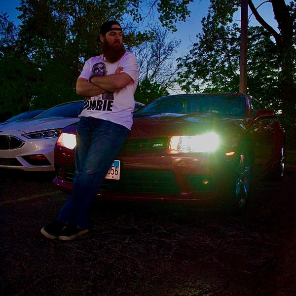 Man leaning on a red car with headlights on at dusk.