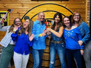 Group of six women smiling and holding axes in a wooden axe-throwing venue.