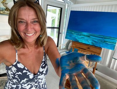 Judy smiling in front of a painting with her hand covered in blue paint reaching out to the viewer,