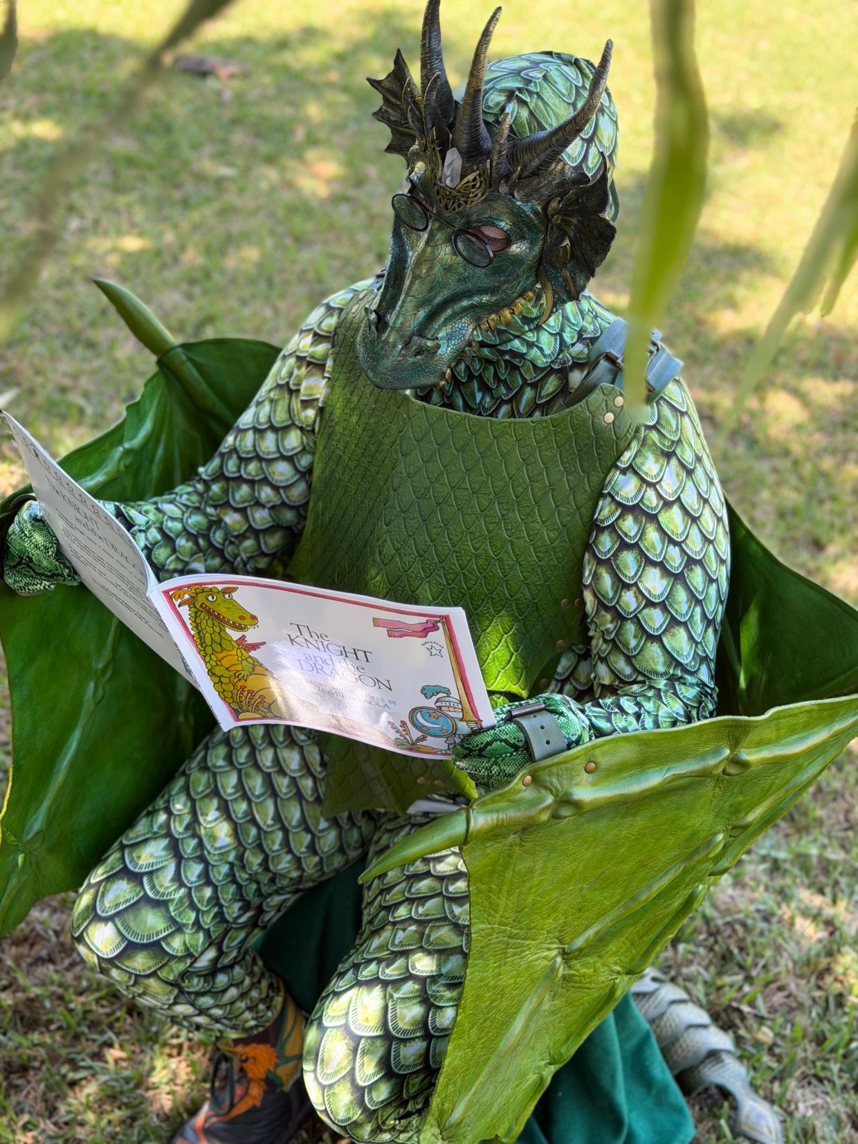 Person in elaborate dragon costume reading a book outdoors.