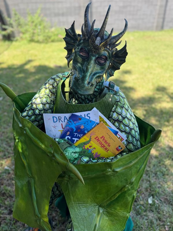 Person in a detailed dragon costume holding children's books outdoors.