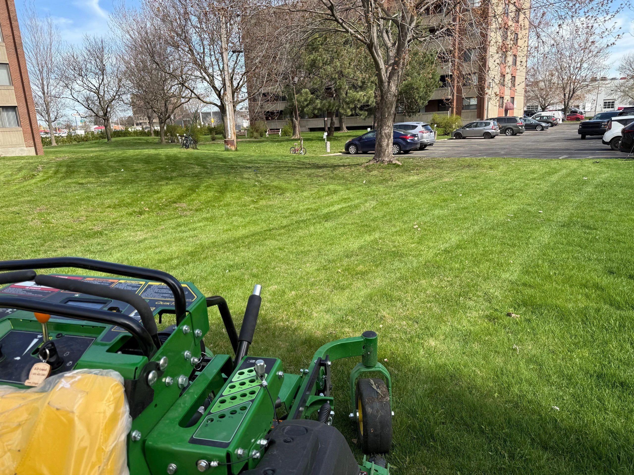 View from a lawn mower cutting grass near a parking lot and trees.
