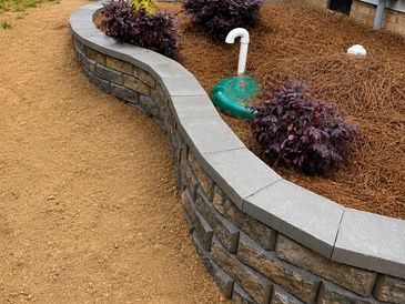 Curved stone retaining wall with mulch and plants in a garden bed.